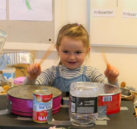 Et barn sidder ved et bord med forskellige objekter arrangeret som et trommesæt. Barnet holder trommestikker og spiller på de improviserede trommer, som inkluderer dåser, plastbeholdere og en metal skål.