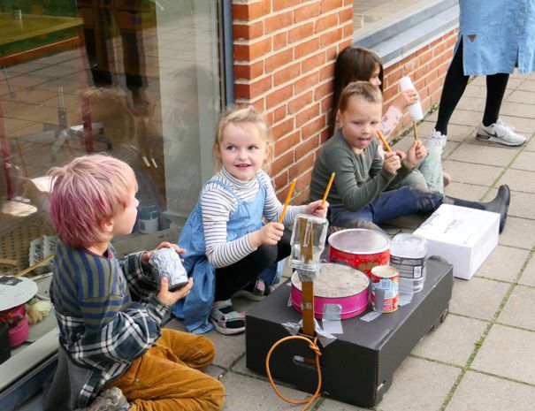 En gruppe børn sidder på fortovet ved siden af en murstensbygning og spiller musik på hjemmelavede instrumenter bestående af forskellige objekter som dåser og kasser.