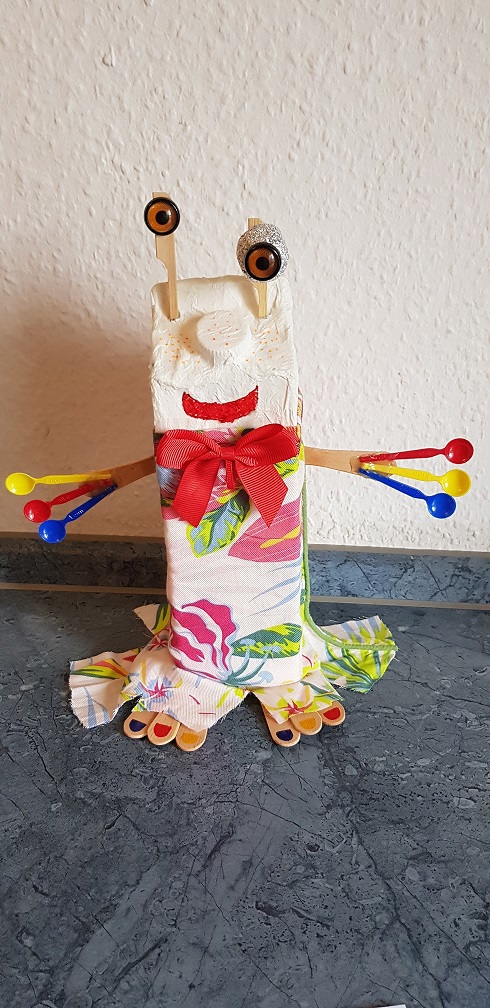 En skulptur af et væsen lavet af en papkasse med blomsterprint, store øjne, en rød sløjfe og farverige skeer som hænder.