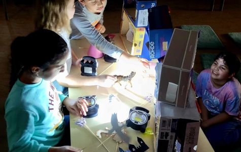 Children experimenting with light and shadows using various objects on a table. A bright light source casts shadows of the objects.