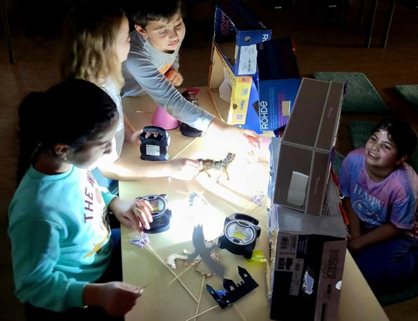 Børn eksperimenterer med lys og skygger ved hjælp af forskellige objekter på et bord. En kraftig lyskilde kaster skygger af objekterne.