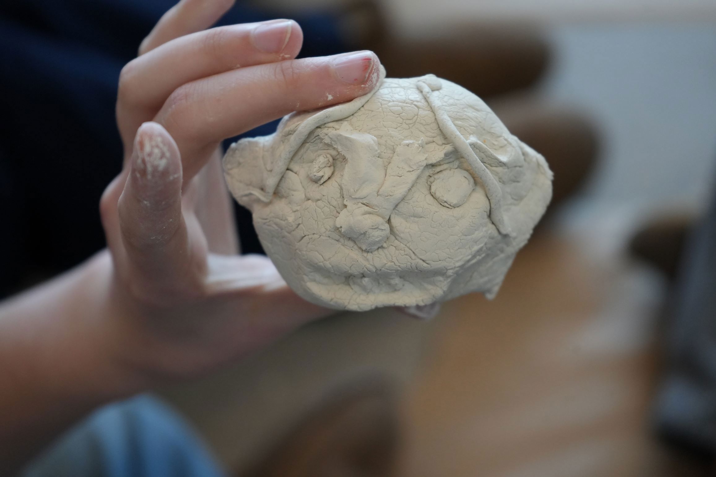 En hånd holder en lille lerfigur af et ansigt med tydelige ansigtstræk, herunder øjne, næse og hårstrå.