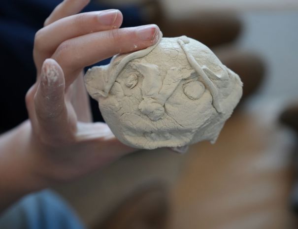 En hånd holder en lille lerfigur af et ansigt med tydelige ansigtstræk, herunder øjne, næse og hårstrå.