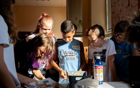 En gruppe børn er samlet omkring et bord med forskellige genstande, herunder papirer.