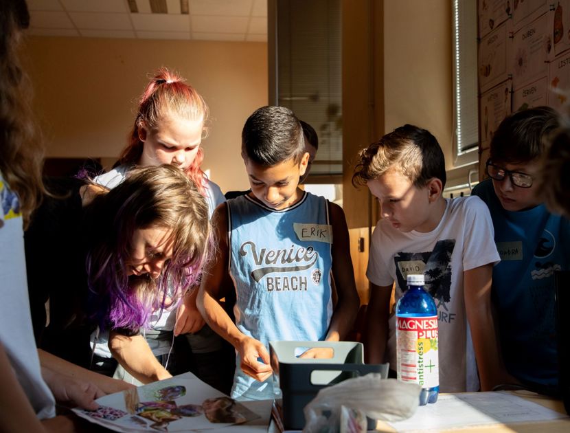 En gruppe børn er samlet omkring et bord med forskellige genstande, herunder papirer.