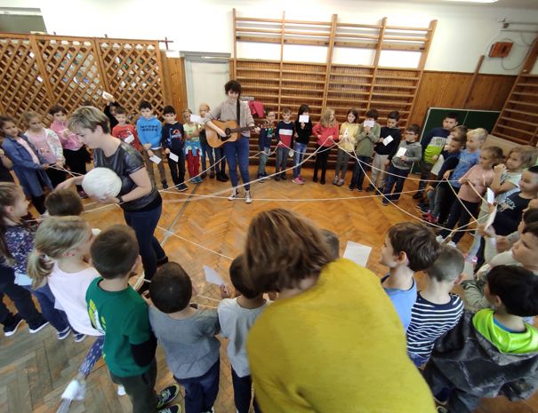 En gruppe børn står i en cirkel og holder snore, der danner en netlignende struktur. En voksen spiller guitar i midten af cirklen, og to andre voksne hjælper med aktiviteten.