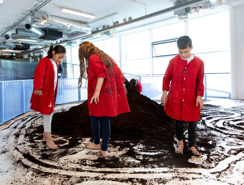 Tre børn iført røde frakker står barfodet på et gulv dækket med jord. De er inde i et rum med industrielle lofter og store vinduer. Børnene interagerer med jorden, som er spredt ud i cirkulære mønstre på gulvet. Der er en stor bunke jord i midten af rummet.