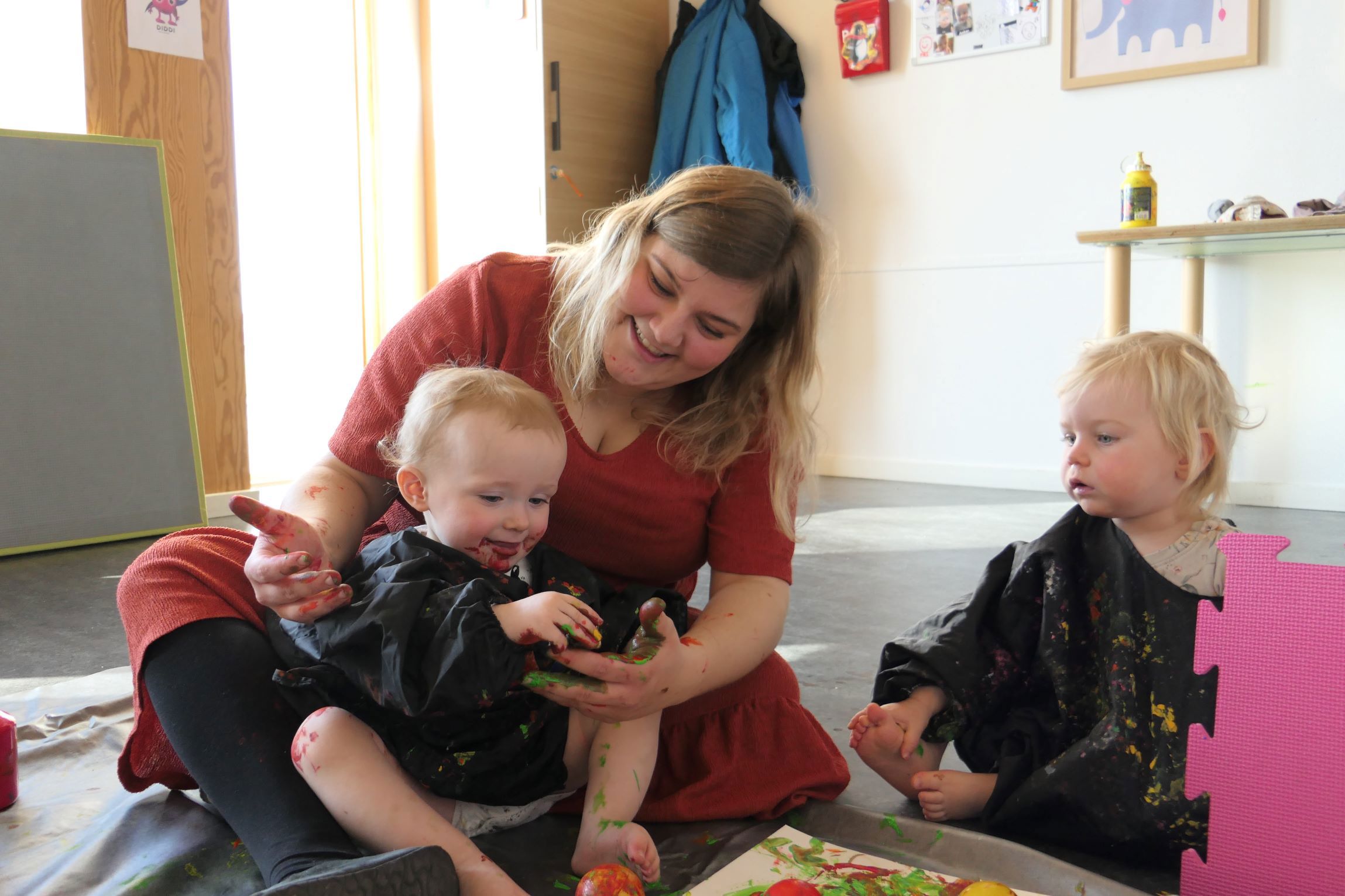 En voksen og to børn, der maler sammen. Den voksne sidder på gulvet med et barn på skødet, mens det andet barn sidder i nærheden. Der er forskellige kunstmaterialer omkring dem, herunder malingsflasker og et lærred med farverig maling.