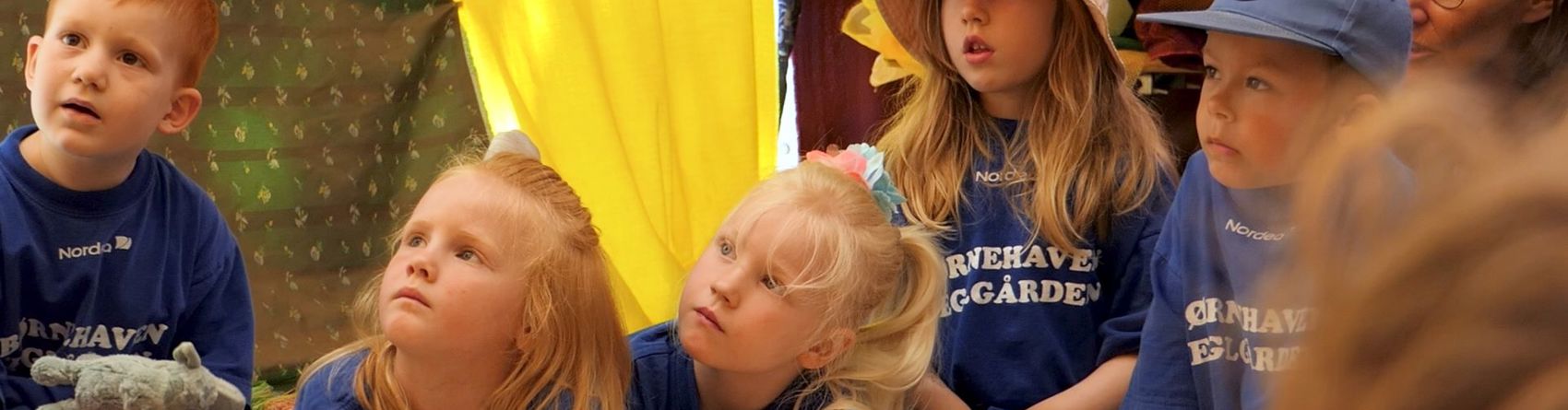 En gruppe børn sidder på en blød, hvid overflade, muligvis et tæppe eller en dyne. Nogle af børnene har også hatte på. Der er en gul og grøn baggrund bag dem.