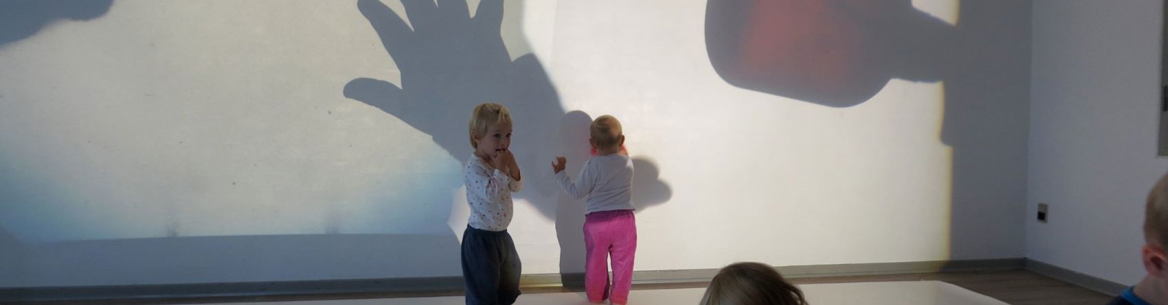 En gruppe små børn i et rum med skygger kastet på væggen. Skyggerne inkluderer en hånd, et fly og en stor genstand, der ligner en flaske eller kop.