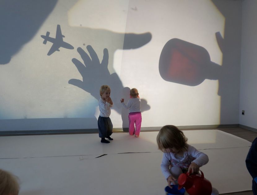 En gruppe små børn i et rum med skygger kastet på væggen. Skyggerne inkluderer en hånd, et fly og en stor genstand, der ligner en flaske eller kop.