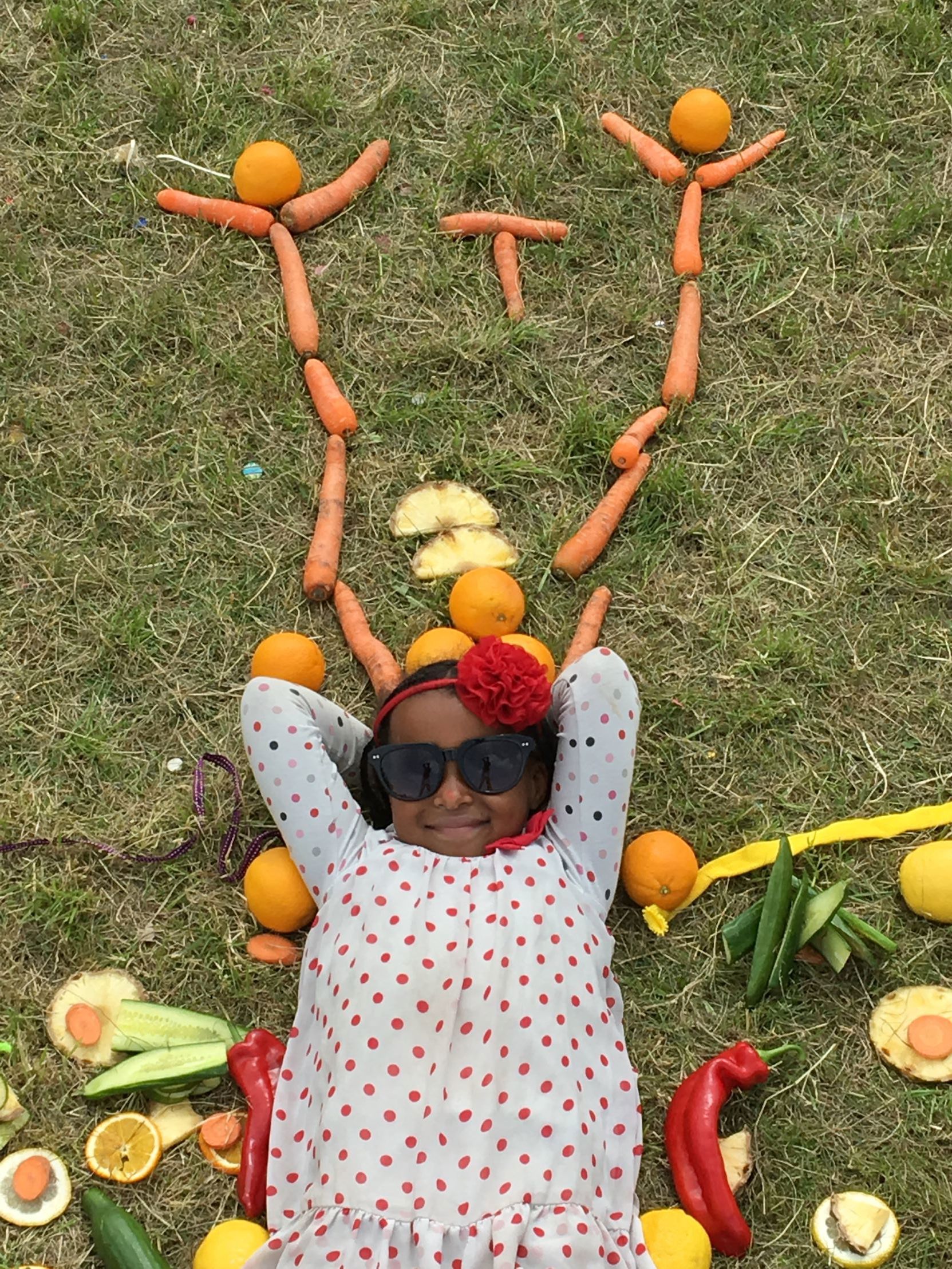 Et barn ligger på græsset med armene bag hovedet, iført solbriller og en prikket kjole. Omkring barnet er der forskellige frugter og grøntsager arrangeret i et mønster, herunder appelsiner, gulerødder, majs, peberfrugter og ananas skiver.
