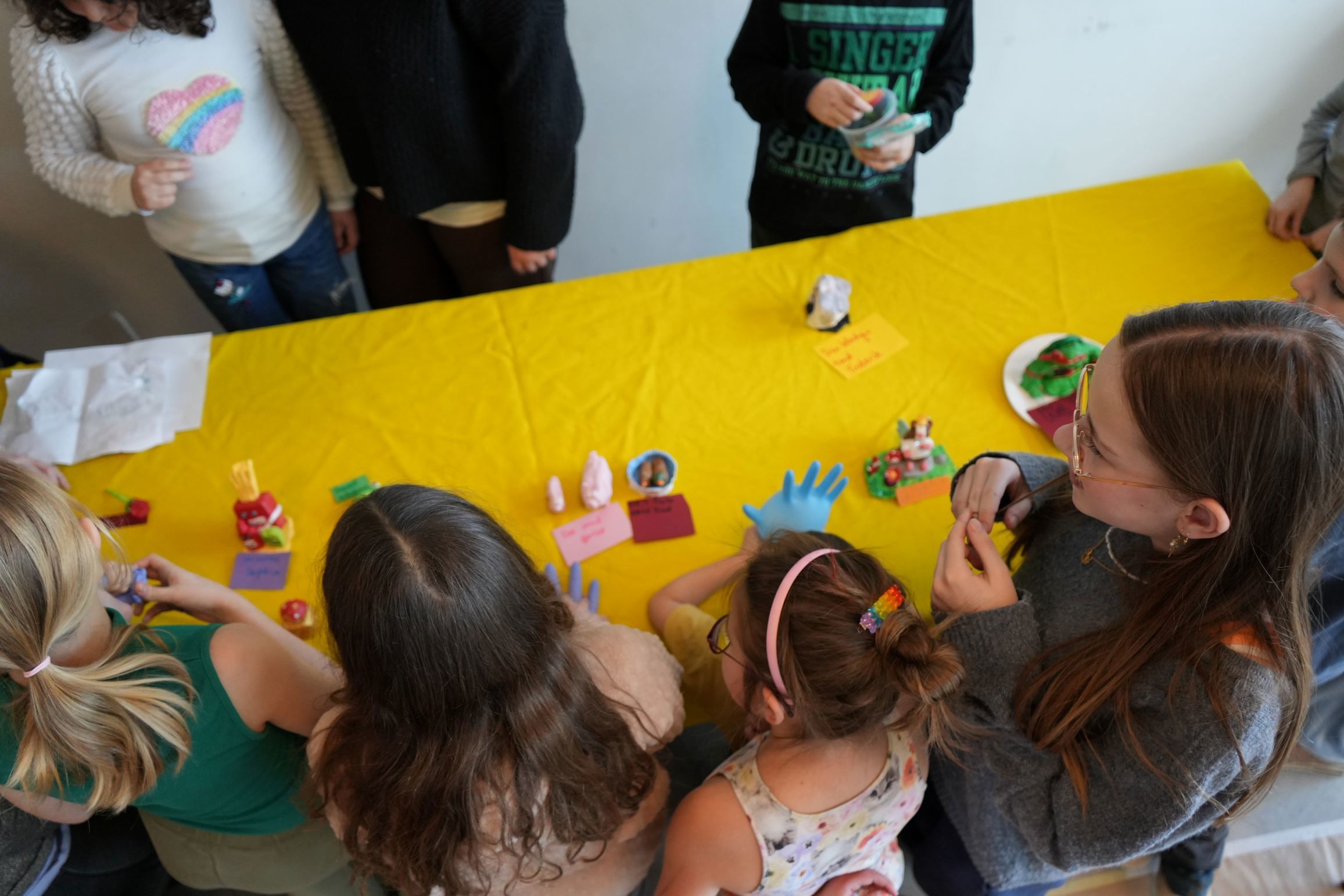 En gruppe børn samlet omkring et gult bord, hvor de viser små skulpturer, de har lavet. Der er forskellige ting og kort placeret på bordet.