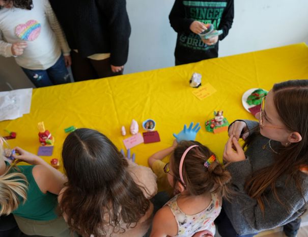 En gruppe børn samlet omkring et gult bord, hvor de viser små skulpturer, de har lavet. Der er forskellige ting og kort placeret på bordet.