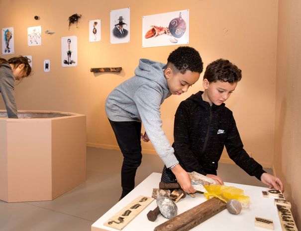 To børn interagerer med objekter på et bord i en udstilling, med billeder og tegninger i baggrunden.