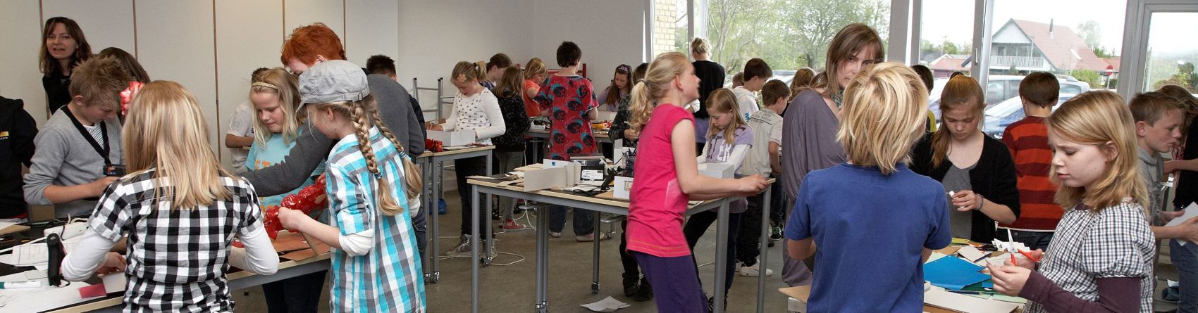 En klasse med mange elever engageret i forskellige aktiviteter. Eleverne står omkring borde og arbejder på forskellige opgaver. Der er flere borde med materialer spredt ud på dem, og eleverne ser ud til at samarbejde i grupper.