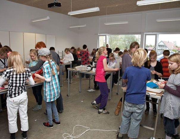 En klasse med mange elever engageret i forskellige aktiviteter. Eleverne står omkring borde og arbejder på forskellige opgaver. Der er flere borde med materialer spredt ud på dem, og eleverne ser ud til at samarbejde i grupper.
