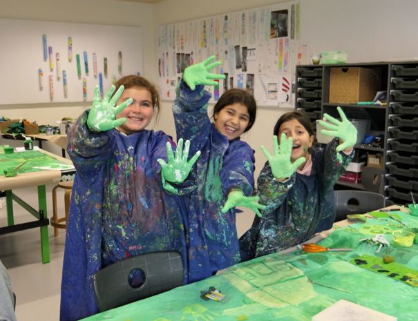 En gruppe børn i et klasseværelse eller kunststudie, iført malerplettede kitler og med grøn maling på hænderne.