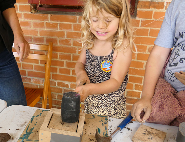 Et barn med langt blondt hår iført et leopardmønstret outfit holder en stor sort hammer. Barnet står ved et bord med forskellige værktøjer og materialer, herunder en anden hammer og noget, der ligner ler. Der er to voksne delvist synlige på hver side af barnet.